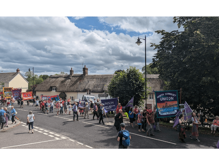 People marching across a village with thatched roofs, holding hand made union banners.