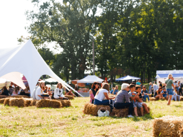 People sat on hay in front of white tents and stalls, an outdoor festival in summer.