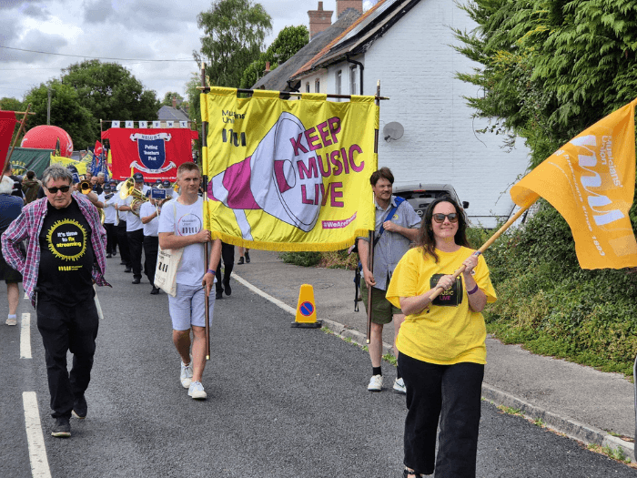 Members and staff marching in the parade waving M U flags, the large bright yellow banner, and wearing t-shirts. Many others are behind them marching in the road.