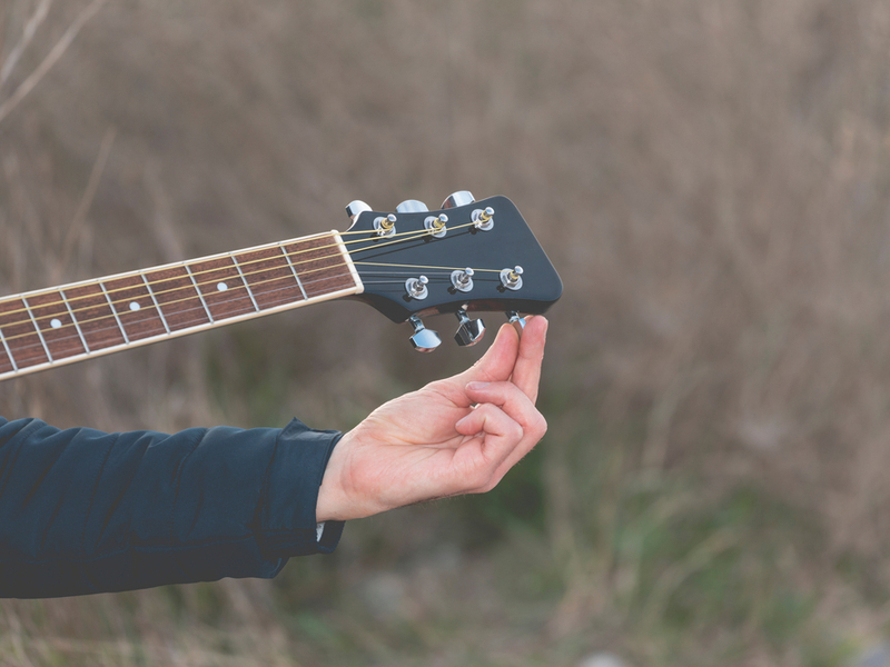 Photograph of a persons hand turning the tuning peg on a guitar, in the background is wild countryside.