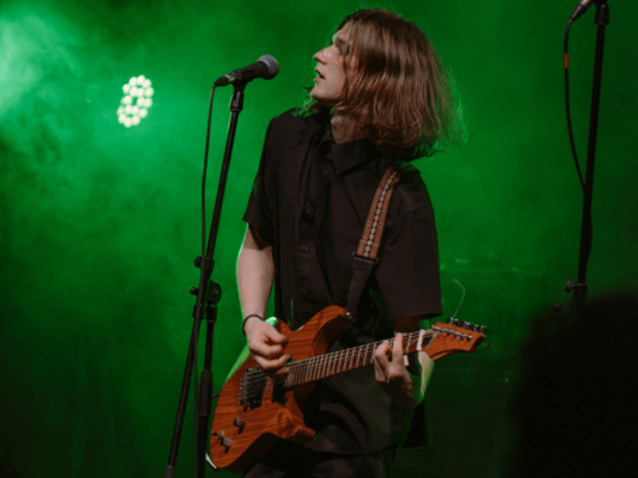 A rock artist with an electric guitar and long hair sings into a microphone on stage. The grunge singer performs at a concert in the spotlight.