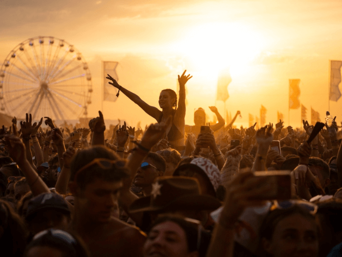 Huge crowds at sunset at Boardmasters Festival, with a ferris wheel and banners in the background. A girl is sat on someone’s shoulders with her hands in the air.