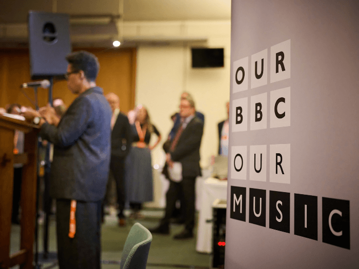 A person at a lectern speaking to a room of people in the background out of focus. In the foreground a sign says 