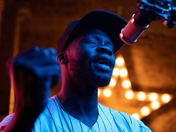 A Black male musician wearing a baseball cap singing into a microphone.