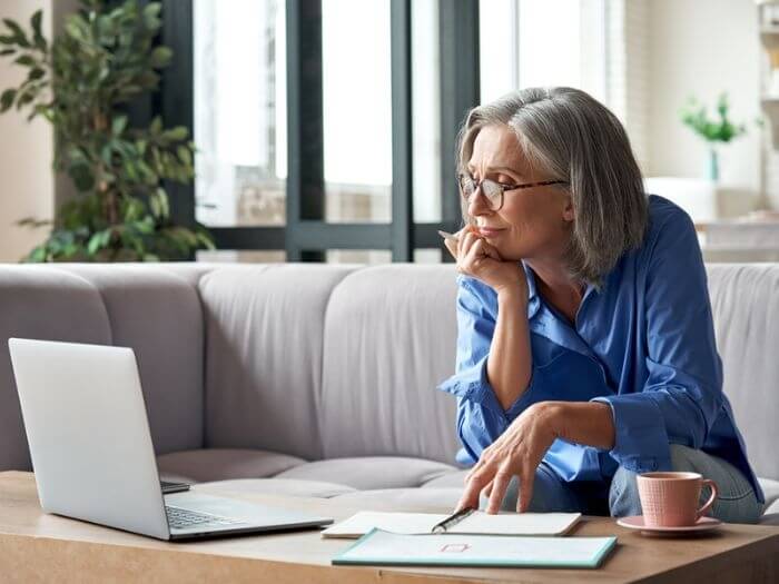 Older woman with glasses sat with a coffee cup and note books in front of an open laptop at home.
