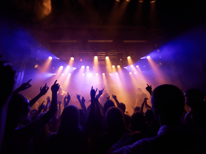 Shot of a large crowd at a small, grassroots music concert. The band on stage are in silhouette against purple, blue and orange lights.