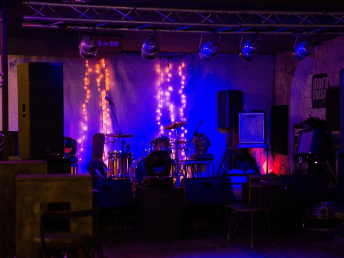 Small, dark, empty stage at a grassroots basement style venue. Instruments are ready and fairy lights line the back wall of the stage.
