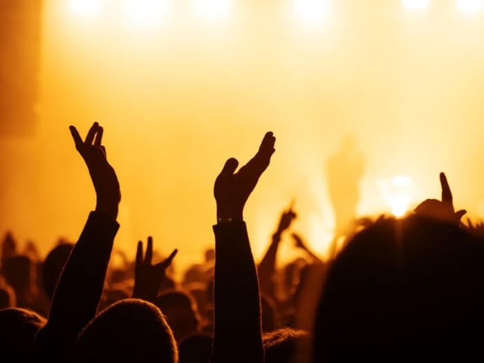 silhouette of raised hands in the crowd at an outdoor music festival, lit by orange and yellow spotlights.