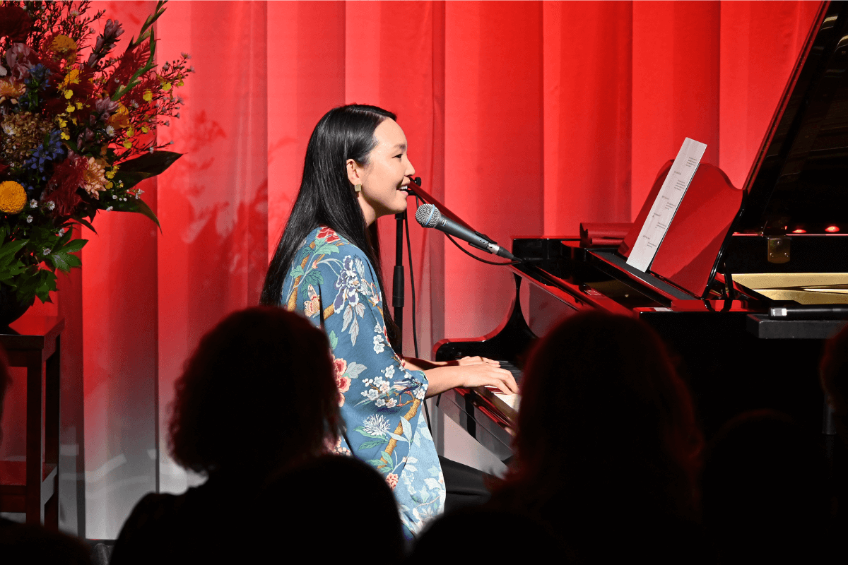 Utako singing and playing piano in front of a crowd, against a red curtain and flowers.