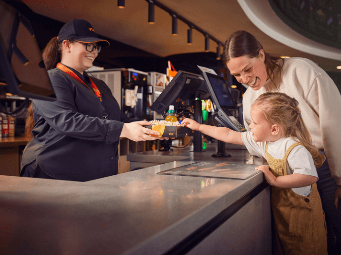 A young child and their parent/guardian smiling as they are handed popcorn across the cinema counter from a staff member working at Vue.