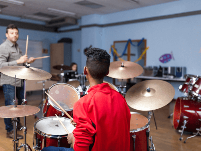 Close up of a child in a red jumper at a drum kit, in a music classroom with other pupils and a tutor.