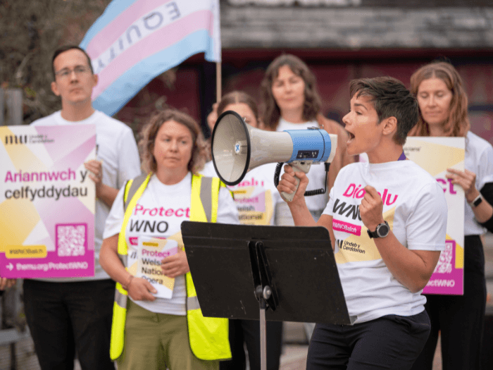 A photo of orchestra musicians wearing campaign t-shirts and holding placards, with one speaking into a megaphone.