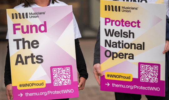 Two people holding placards saying 'Fund The Arts' and 'Protect Welsh National Opera'.