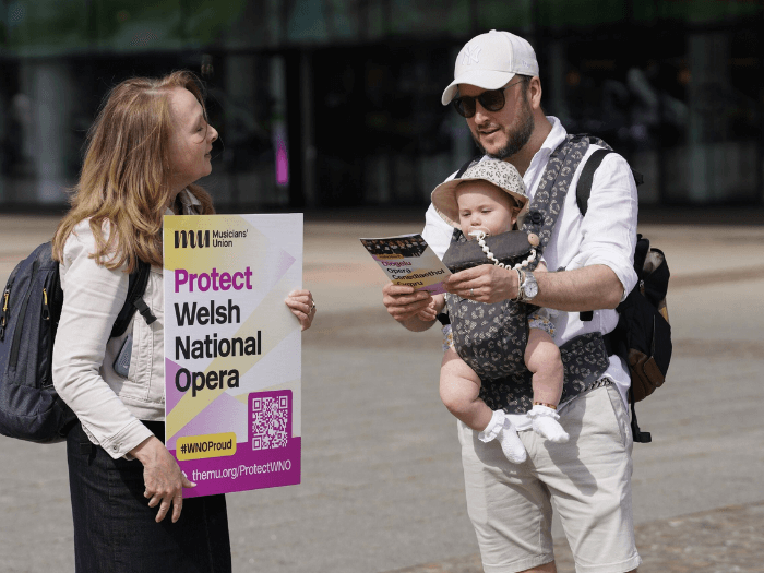 A MU Wales Official holding a sign and handing out flyers to a man and his young baby during a demo outside of The Wales Millennium Centre in June.