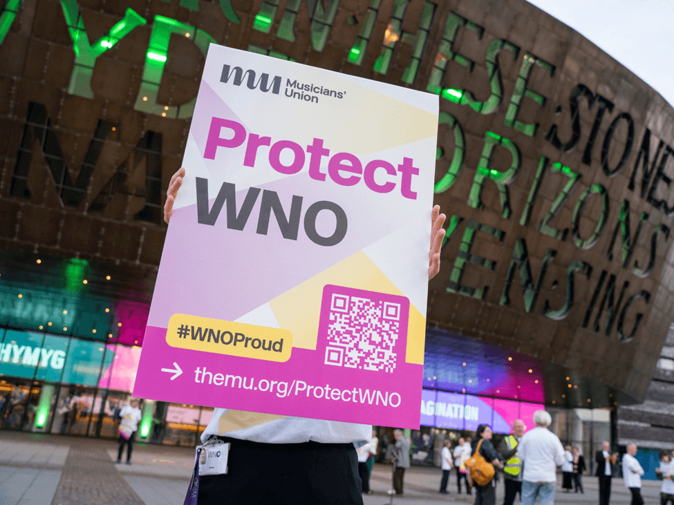 A Welsh National Opera orchestra member holding a campaign placard at a demo.