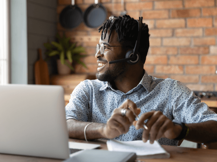 Young Black man with over ear headphones on, smiling holding a pen looking away from an open laptop.