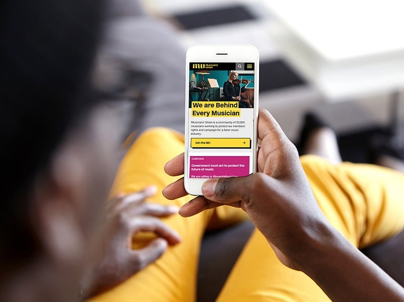 Photograph of hands holding a mobile phone which is displaying a view on the MU's new website's homepage. The person is wearing yellow trousers which are visible in the background.
