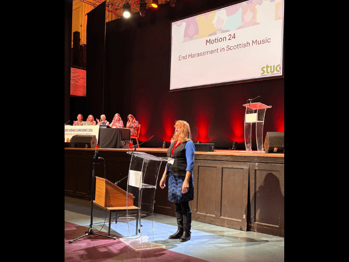 Wendy standing at the front of the room in front of a lectern, behind her is a screen saying 