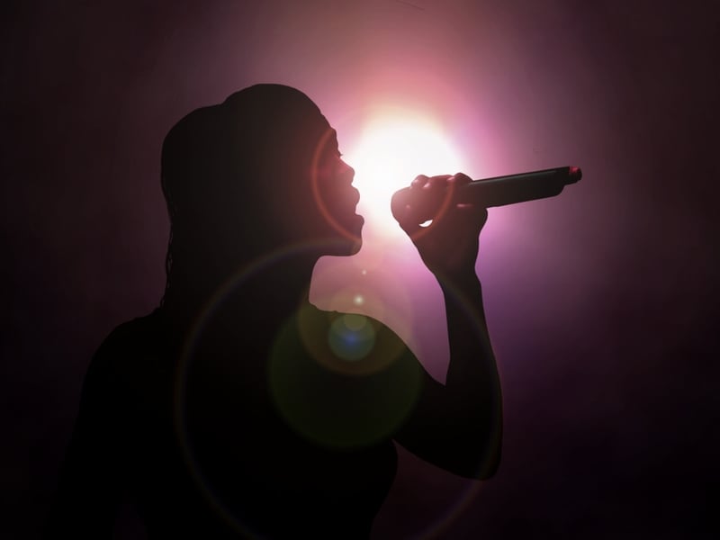 Photograph of a singer holding a microphone up to their face against a purple misty background, the rest of the image is in shadow.