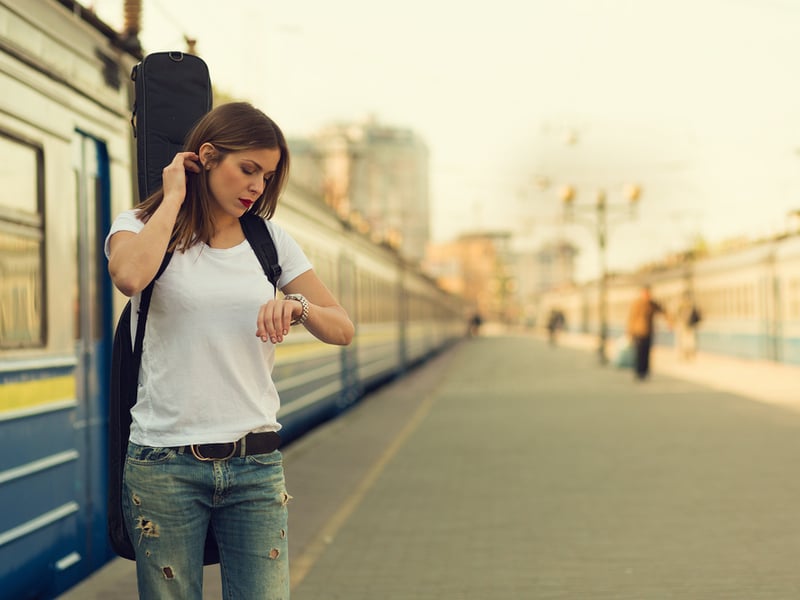 Woman waits by a train with a guitar strapped to her back, she is looking at her watch. Photo credit: Shutterstock