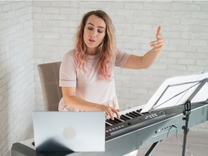 A woman with long blonde hair dyed pink at the tips makes a gesture with her hand while sat an electronic keyboard. A laptop computer is propped up to broadcast from.
