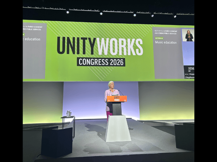 Xenia Horne on stage at a lectern, a green coloured screen behind her says "Unity Works Congress 2026".