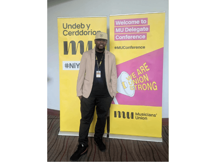 Xidus in front of banners that say 'Welcome to M.U Delegate Conference'.
