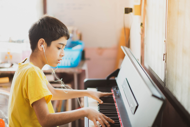 Photograph of a young boy in a yellow t-shirt, playing on an electric piano with earphones in that we assume are helping him learn from a distance.