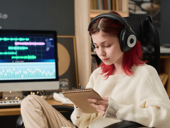 Young female musician with red hair, sat in front of a computer mixing a song, writing in a notepad with headphones on.