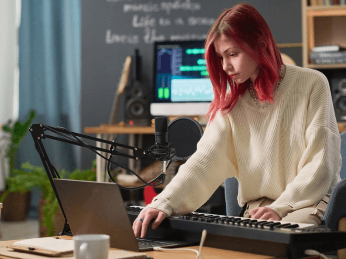 Young adult women with red hair in a recording studio, looking at a laptop while playing keyboard.