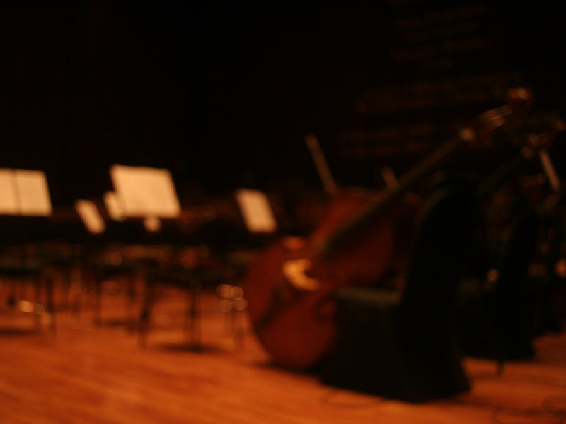 Blurred photograph of an empty orchestral set-up on a stage, we can see empty seats, music stands and an unmanned double bass.