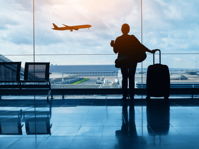 A woman standing in an airport with a suitcase watching a plane take off through the window.