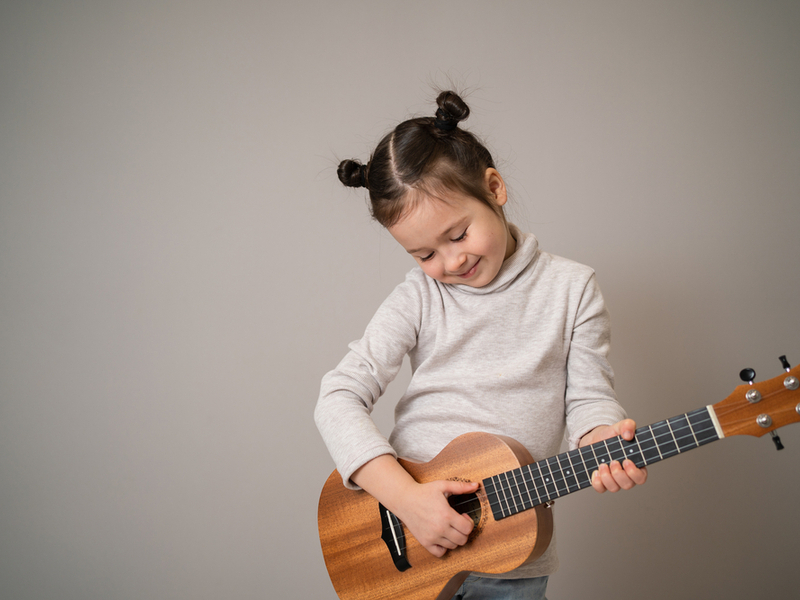 A small child holds a ukulele and smiles fondly downwards against a plain background.