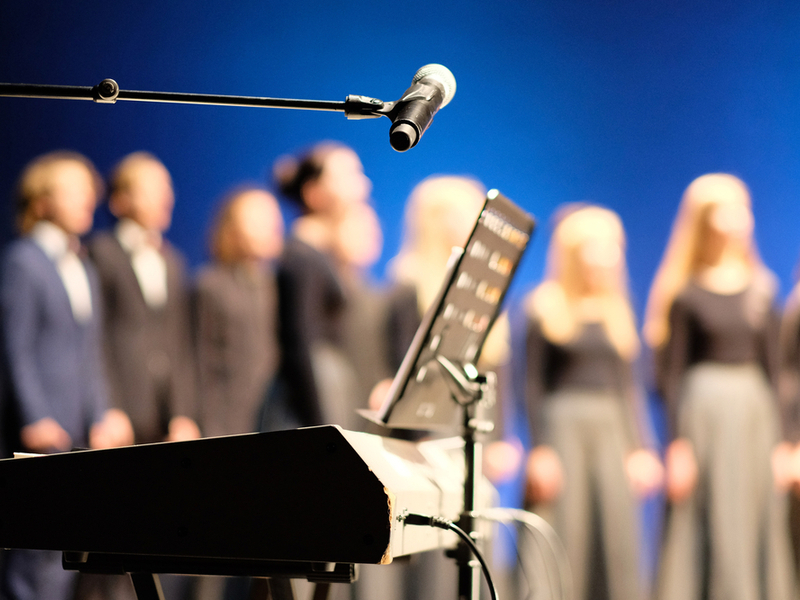 Photograph of a choir, blurred in the background, with a keyboard and microphone in sharp focus.