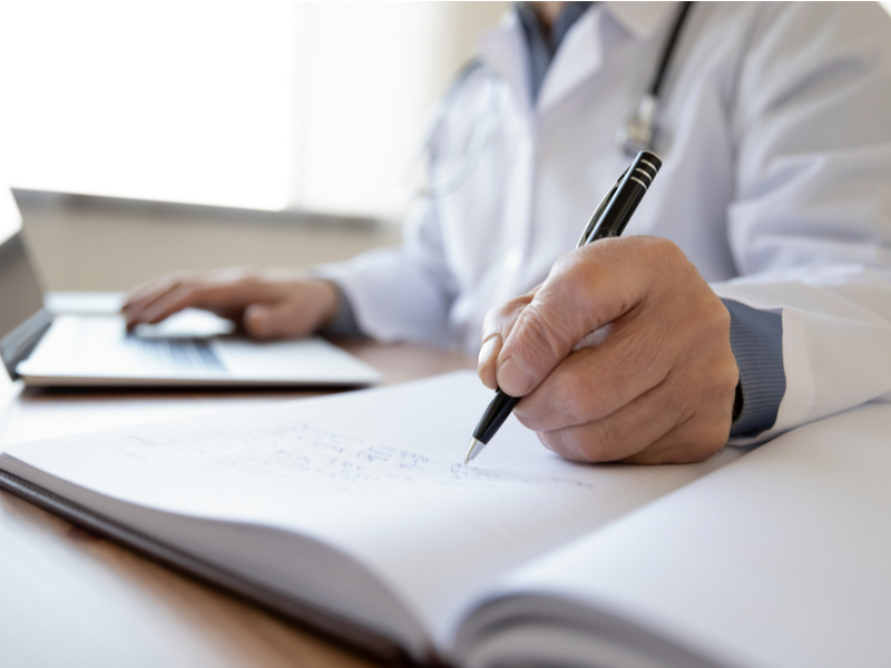 Photograph of someone writing into a notebook wearing a white coat in what appears to be a medical setting