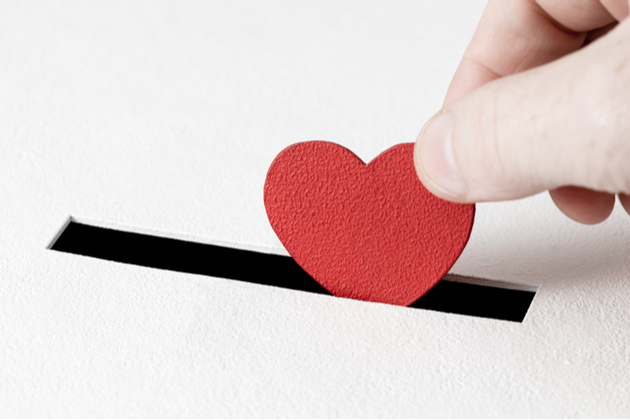 Close up of hand putting a red heart into the slit of a white donation box