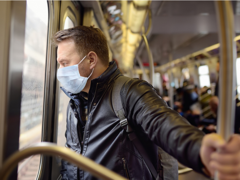 Man wearing blue face mask in leather jacket with short hair, looking out of a window waiting to get off a bus.