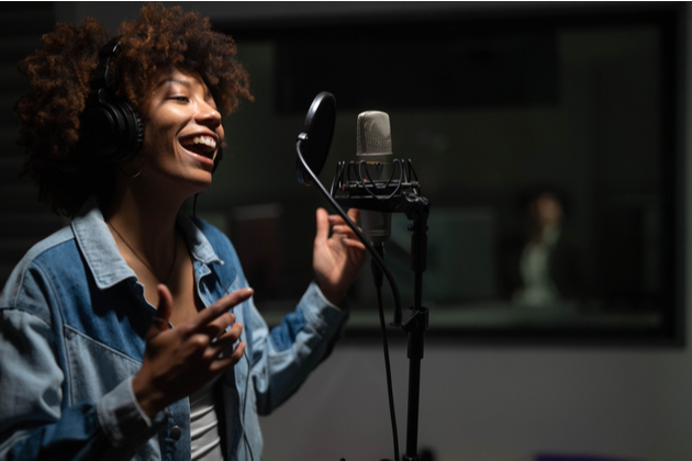 Black female singer wearing headphones is performing and recording a song in a music studio.