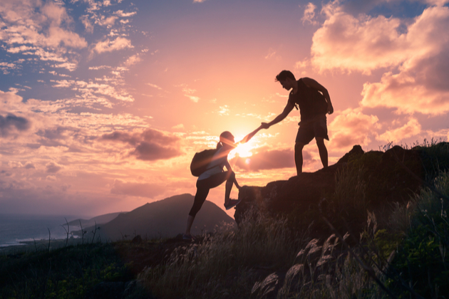 Two climbers set against a sunset on a mountain, one offering the other a hand up.