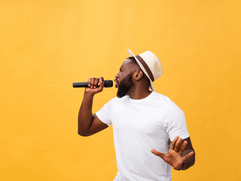 A man is singing with energy into a microphone, against a yellow background.