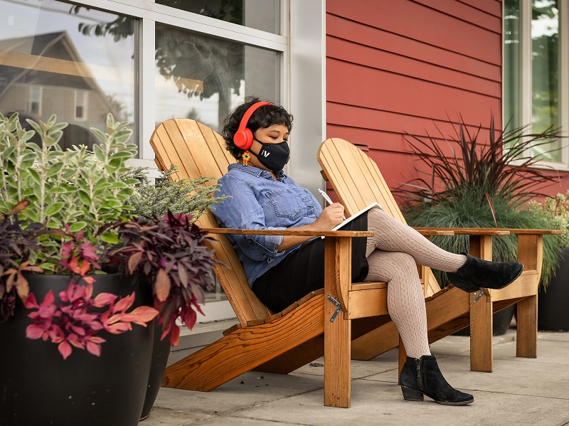 Photograph of a masked woman sitting outdoors, writing into a notebook and wearing headphones.