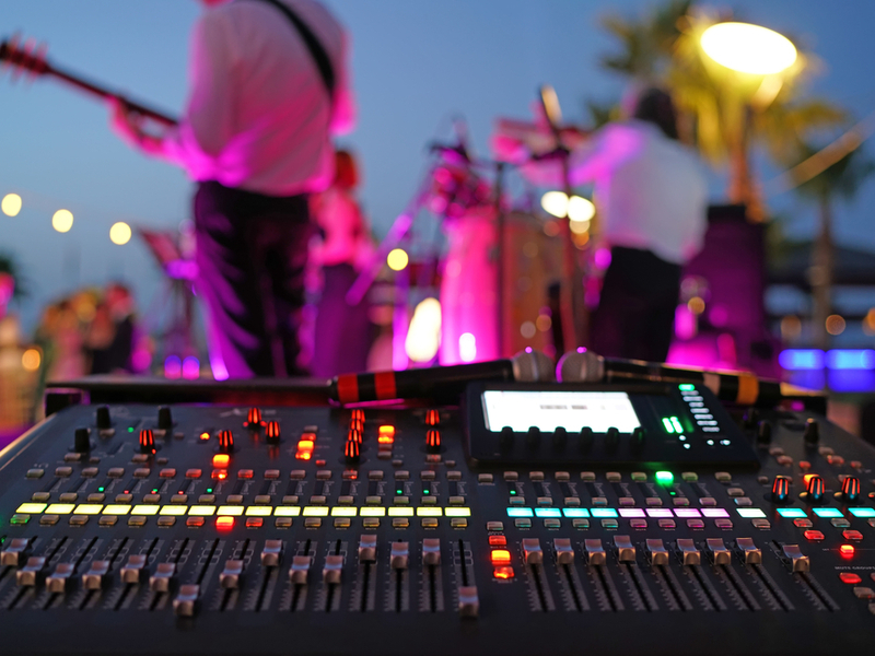 Photograph of a mixing desk, behind which we can see the blurred figures of musicians performing on stage under bright lighting.