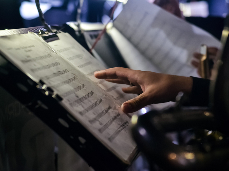 Photograph zooming in on a hand moving to turn the page on a score, blurred in the dark background are further scores in what appears to be an orchestral section.