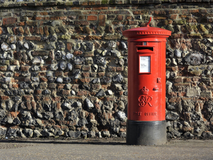 Red post box stands in front of a brick wall in the bright sunshine.