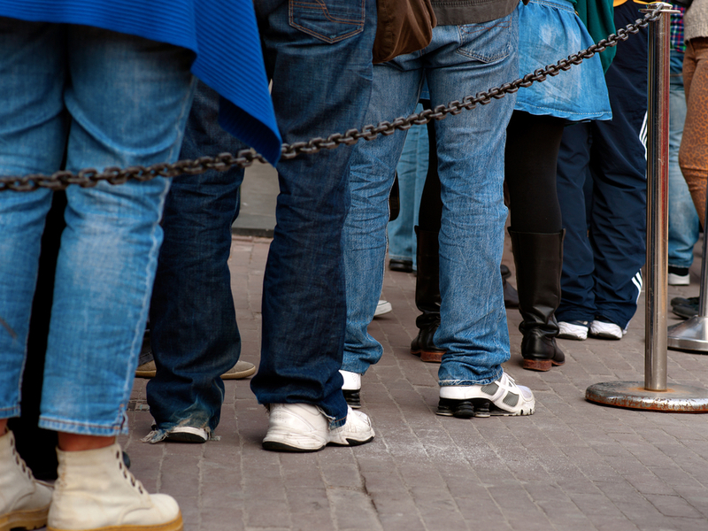Photograph of a number of peoples legs, standing in a queue in the daylight behind a small, temporary fence.