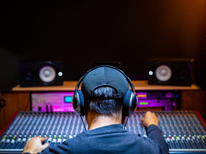 A person is sat at a large recording desk, we can just see the back of their head with short cropped hair, a baseball cap, and earphones over their ears.