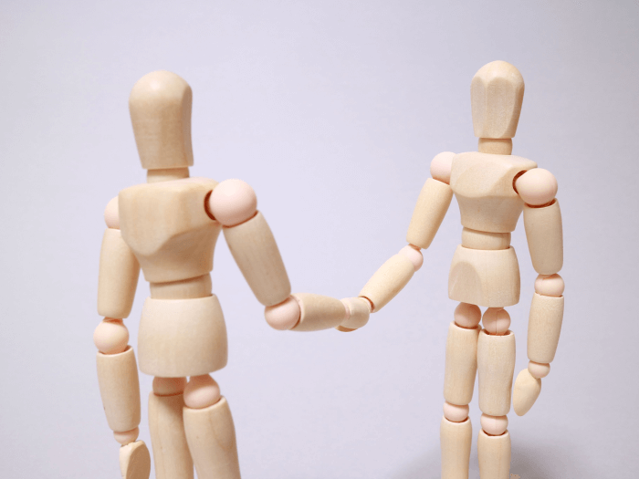 Two wooden dolls shake hands against a pale lilac background