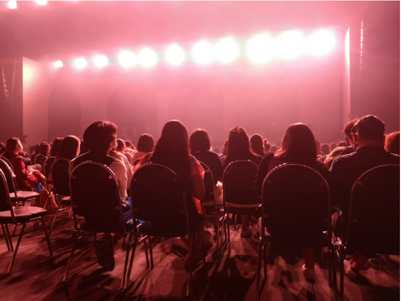 Photograph of a socially distanced audience watching a live performance from the back. We see them from behind, seated on distanced chairs with a red light.