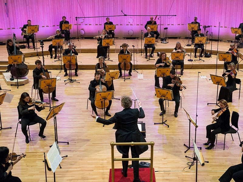 Photograph of an orchestra set out in a socially distanced way, we can see all the musicians sat out across the stage in distanced positions, whilst a conductor gestures from the front.