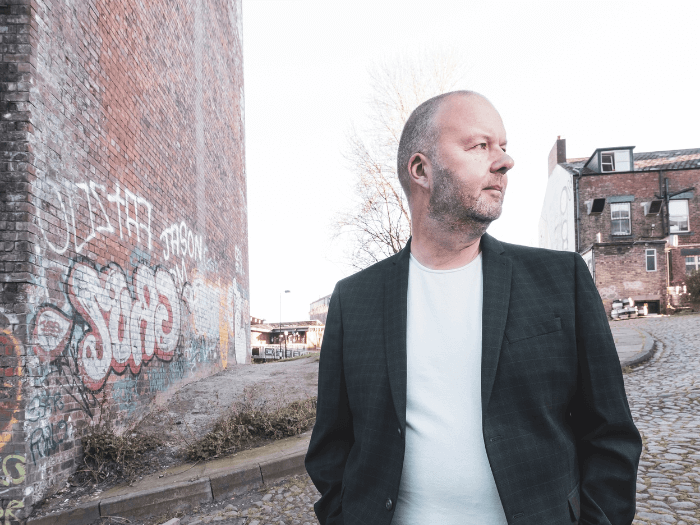 Musician Steve Luck in a white t-shirt and black blazer, standing next a graffitied wall on a cobbled road.